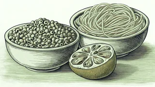 A bowl of black eyed peas and a bowl of noodles stand on a wooden table beside a halved round fruit.